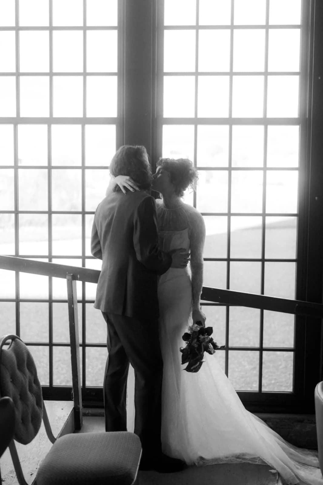 Bride and groom embracing beside tall windows.