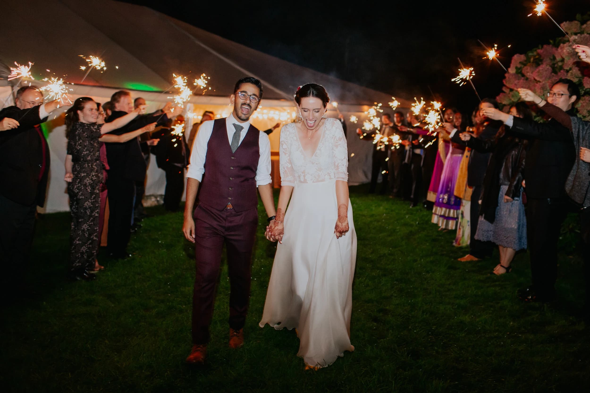 Bride and groom walking through guests holding sparklers at night.
