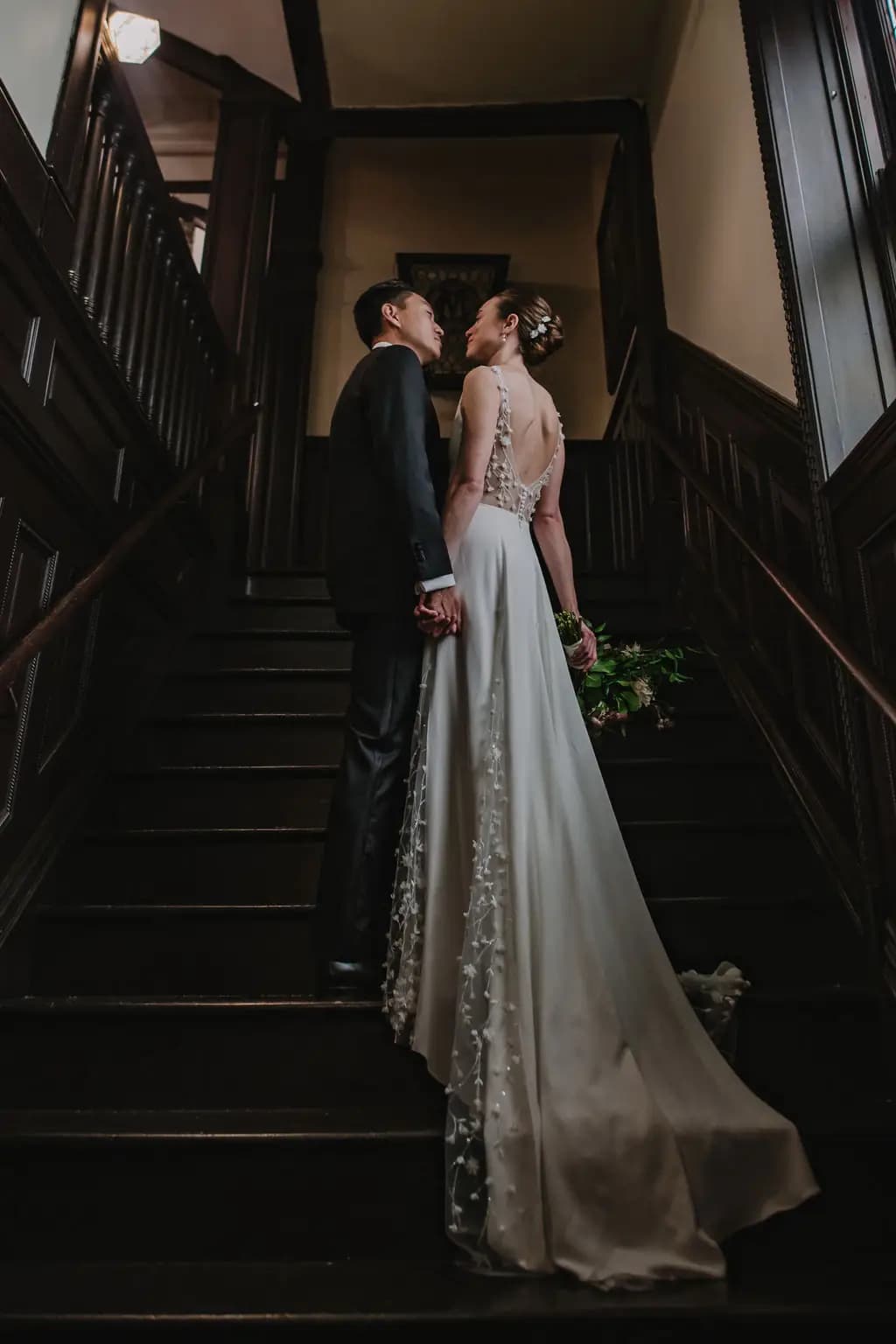 Bride and groom holding hands on a dark wooden staircase.