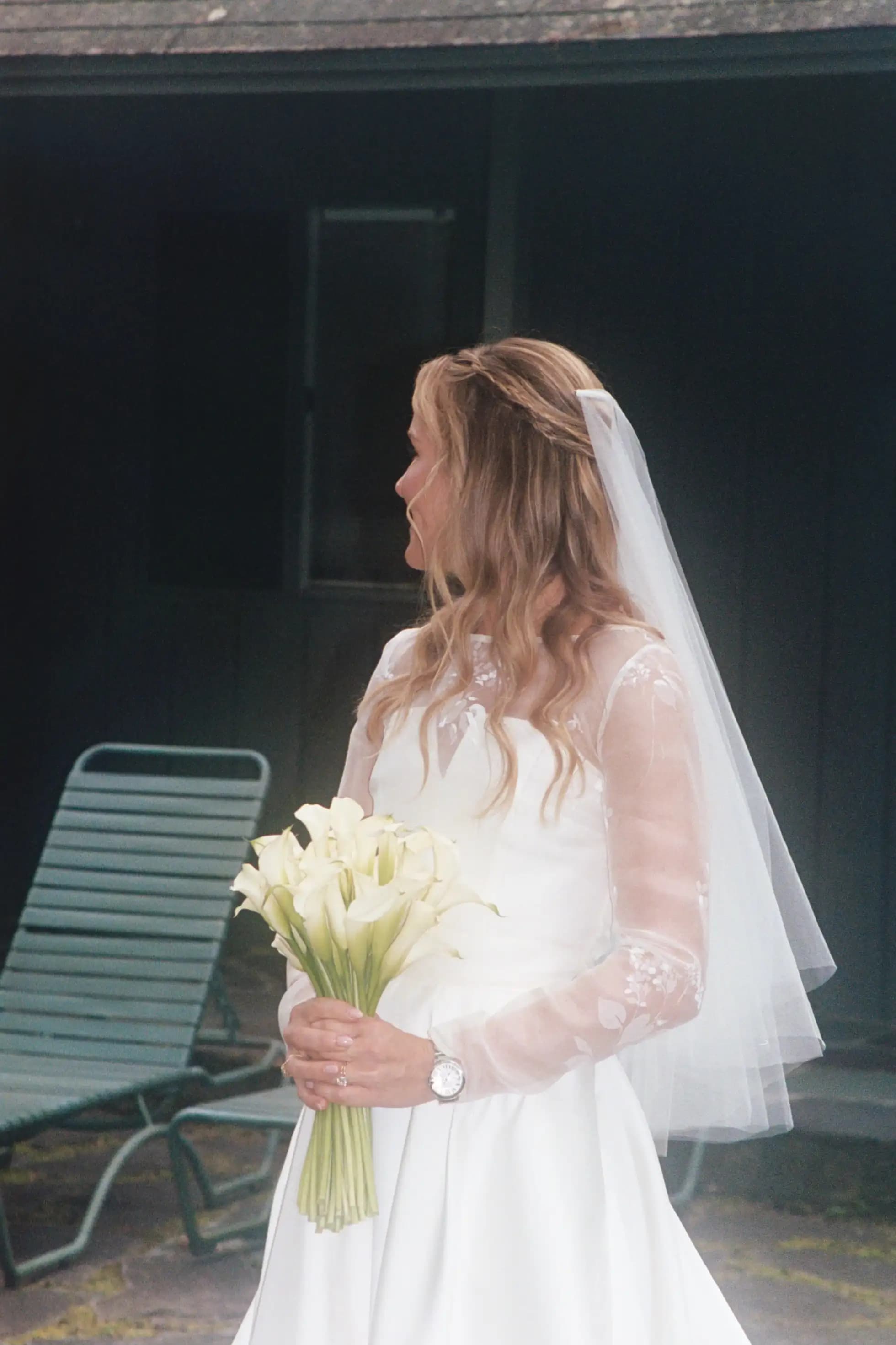 Bride in profile holding white lilies beside a dark doorway.