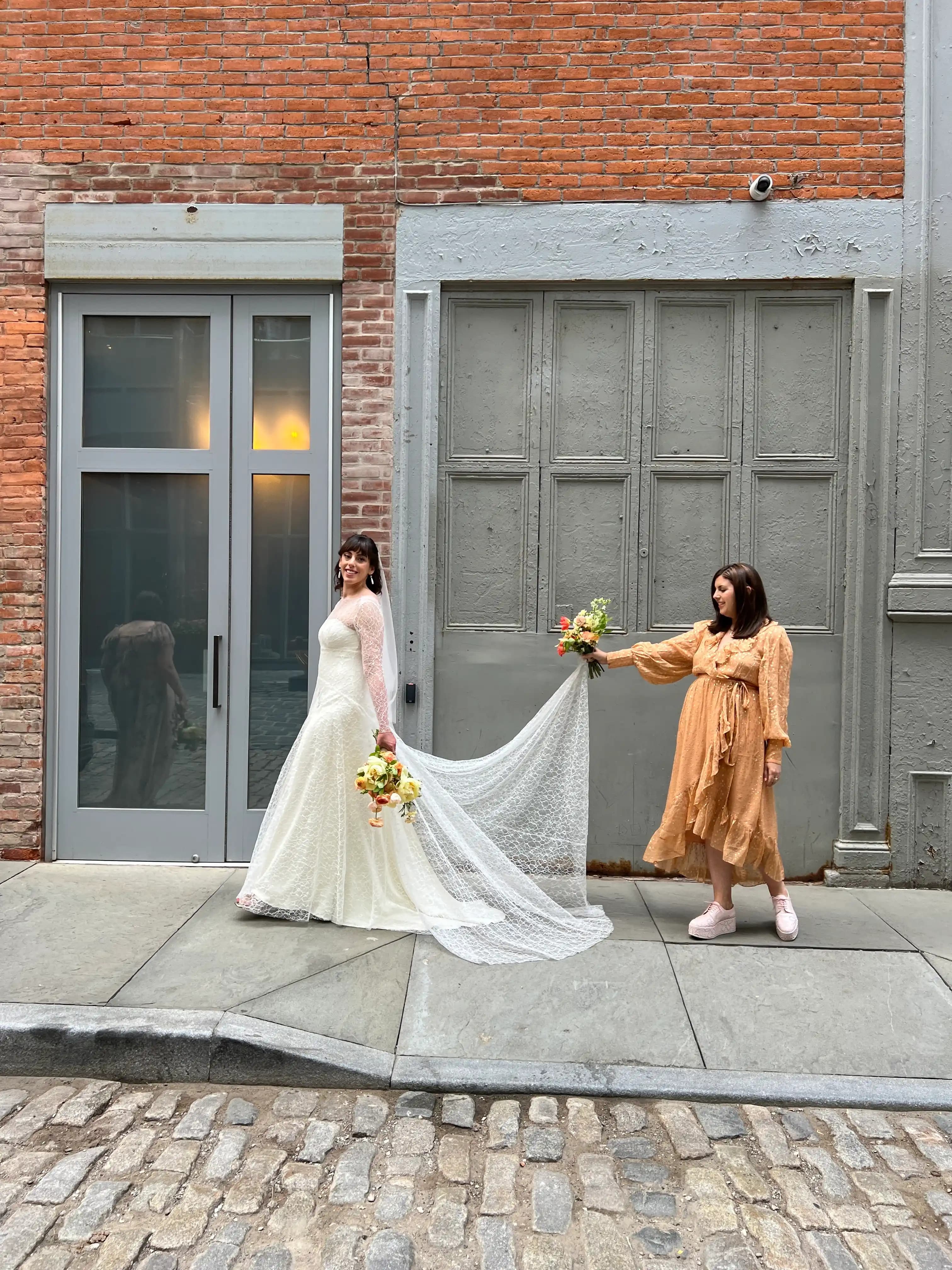 Bride holding her train while a friend carries flowers in a city alley.
