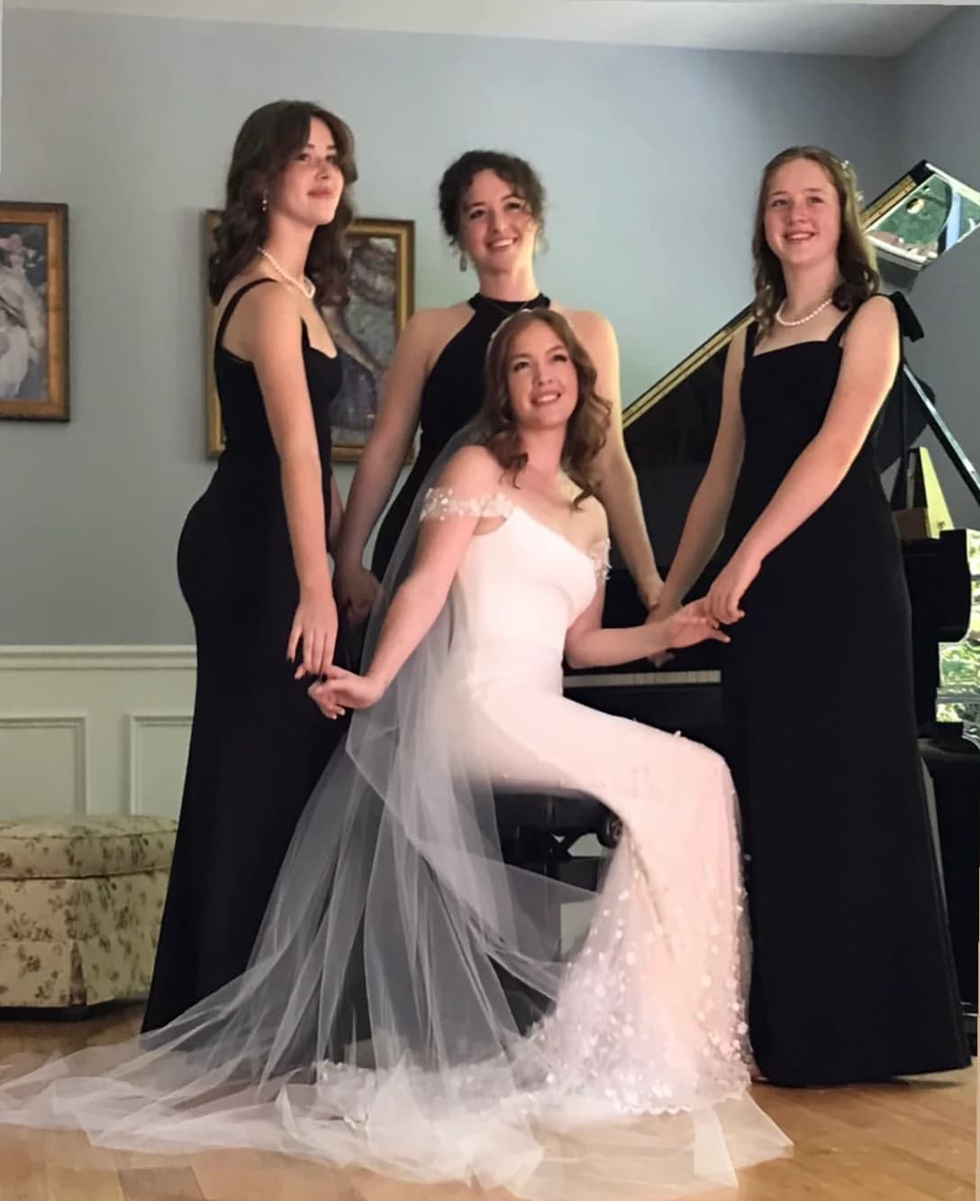 Bride seated with three attendants in black dresses beside a piano.