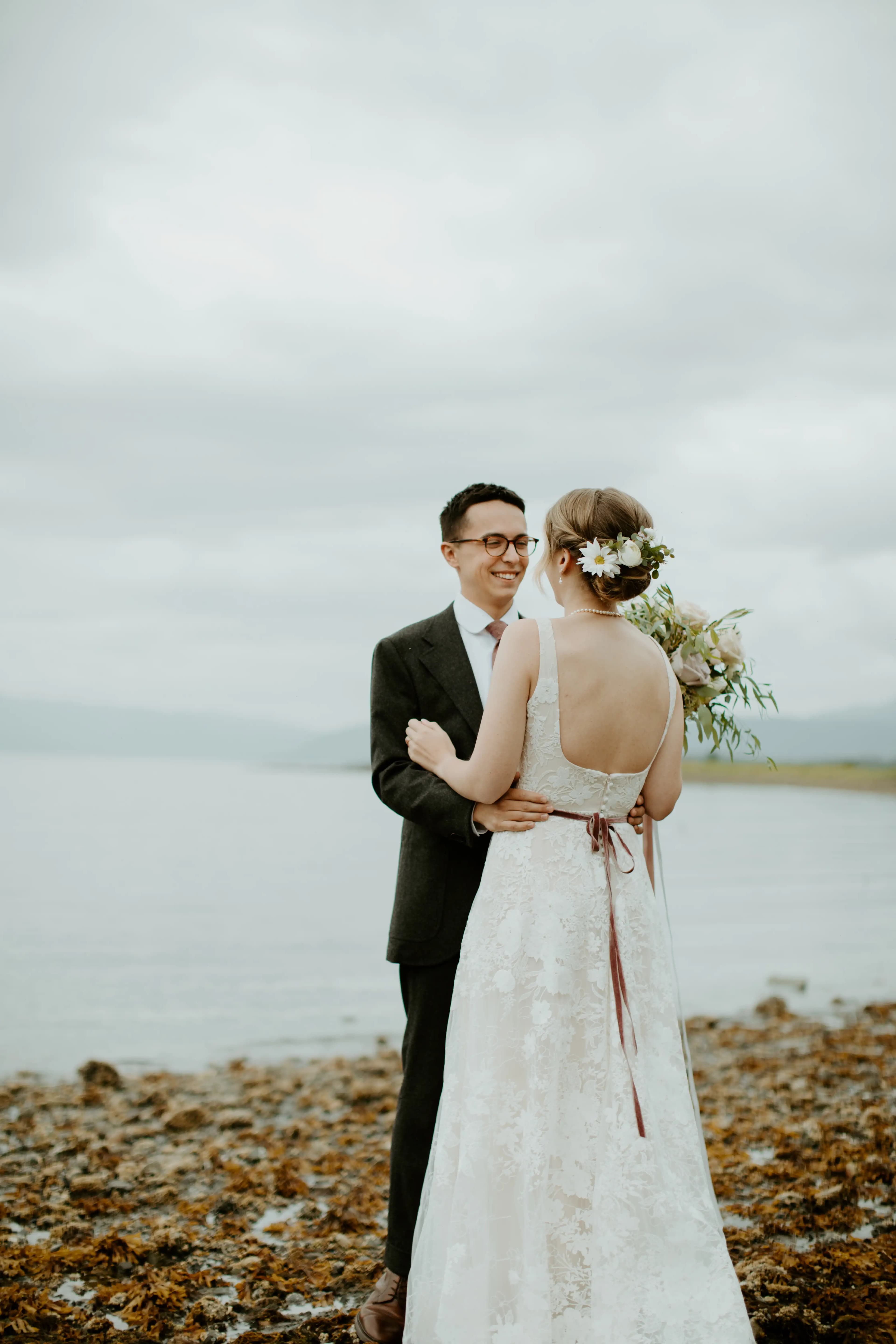 Bride and groom embracing beside a lake, with the bride facing away.