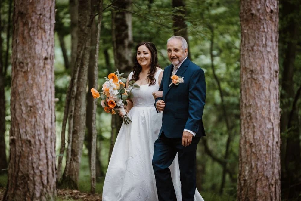 Bride walking through a forest with an older man beside her.