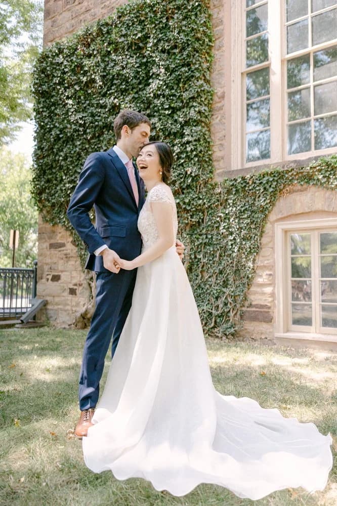 Bride and groom laughing beside an ivy-covered stone building.