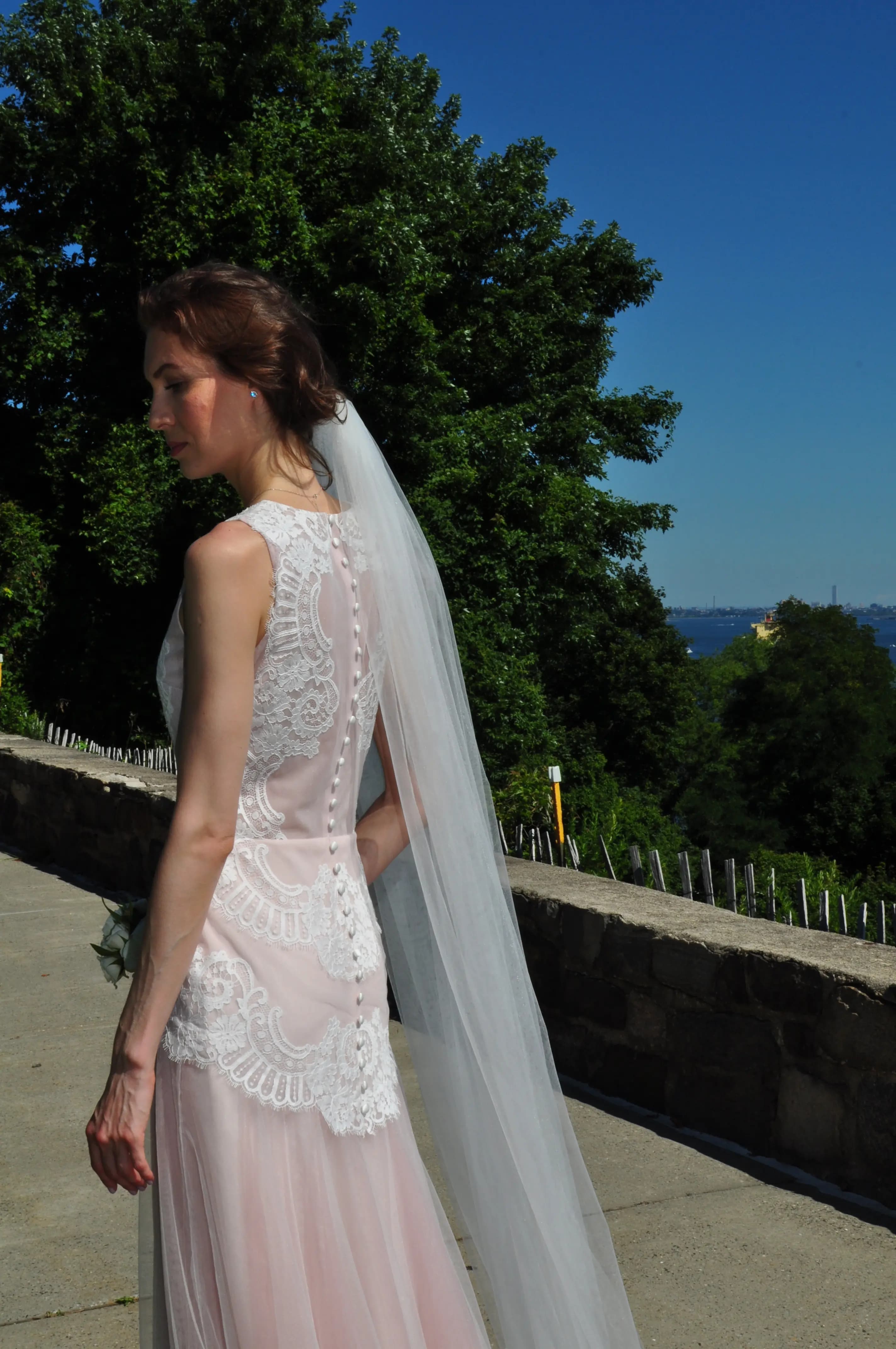 Bride showing the back of a lace gown and veil beside the water.