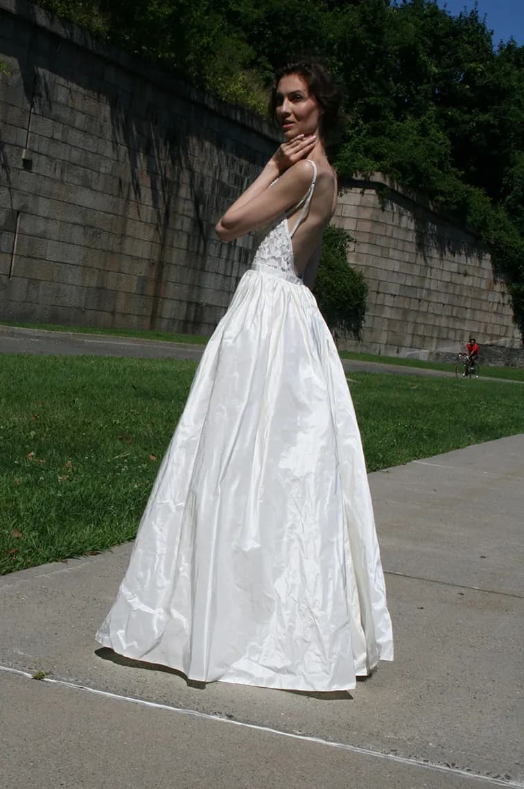 Bride showing the open back of a gown beside a stone wall.
