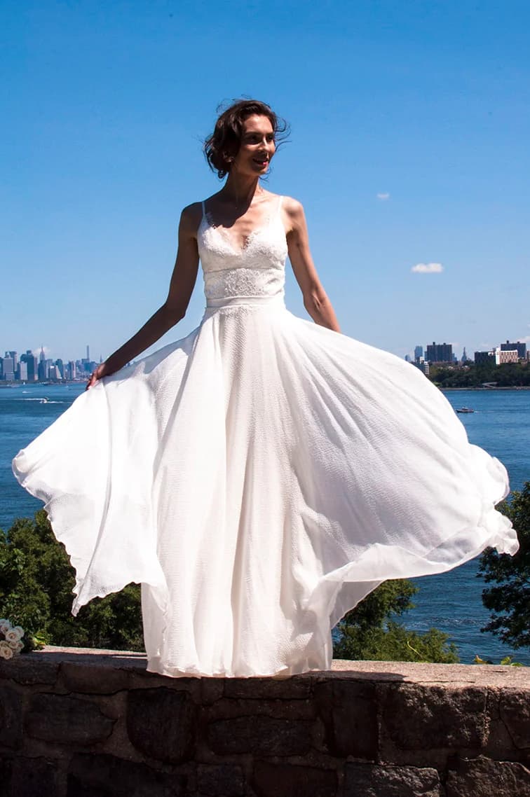 Bride lifting the skirt of a gown beside the water.
