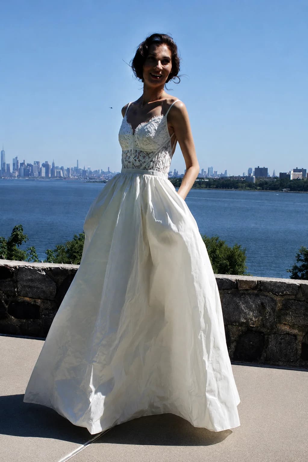 Bride in a full skirt and lace bodice beside the water.