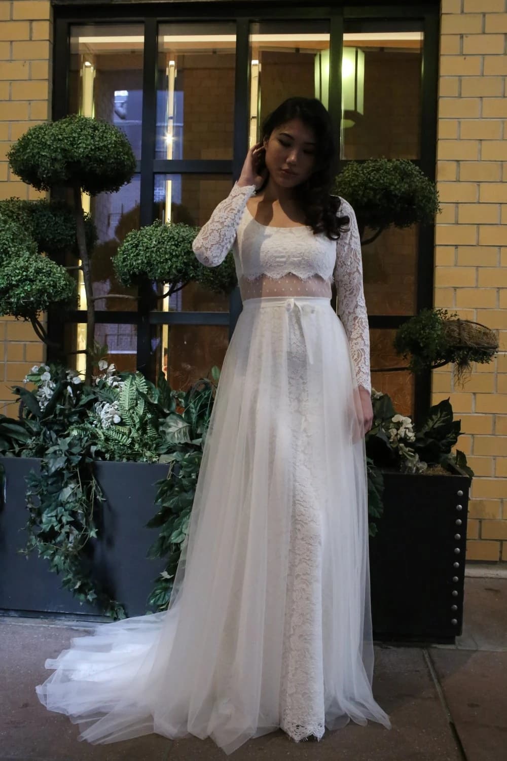 Bride in a lace two-piece gown beside topiary trees.