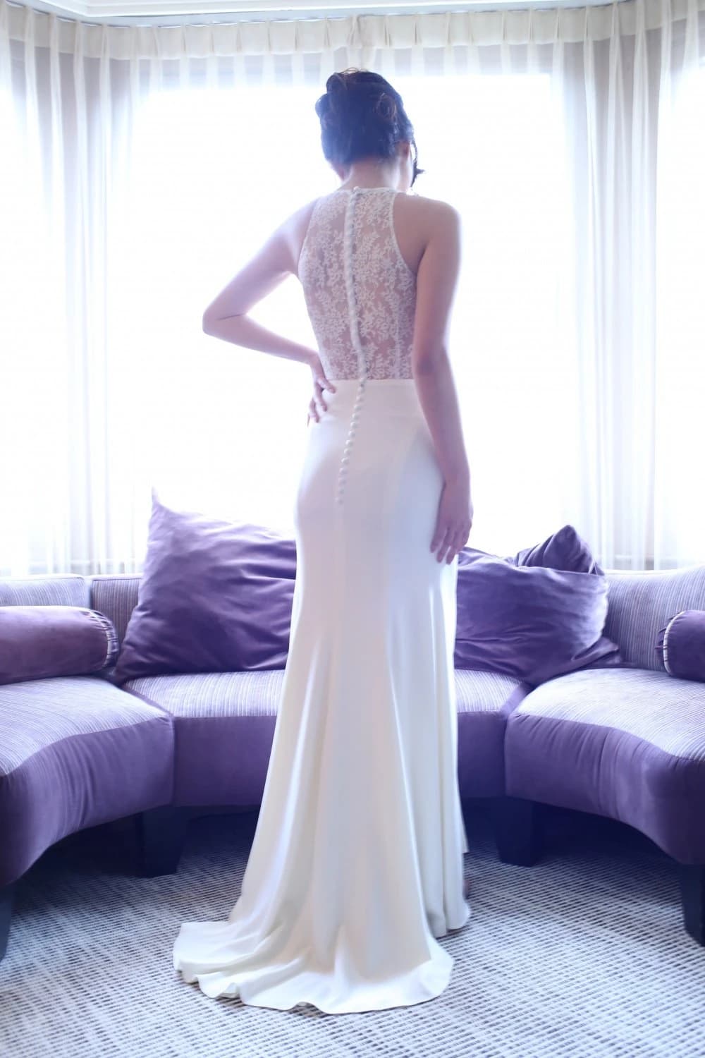 Bride viewed from behind in a lace gown before a window.