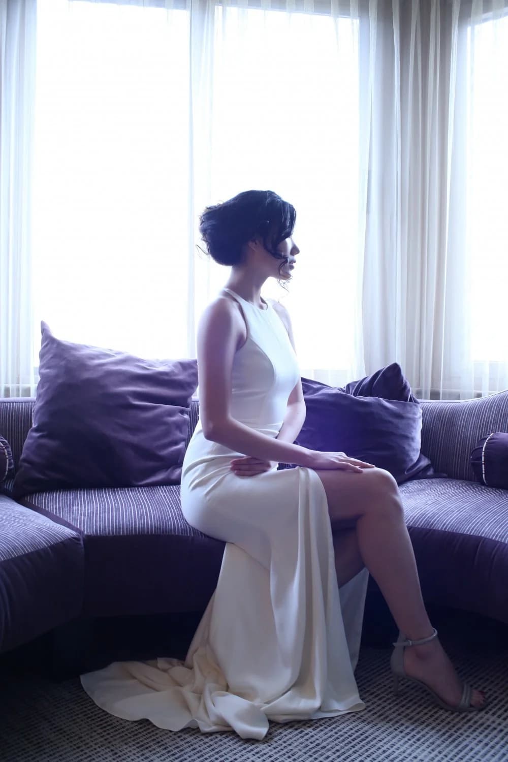 Bride seated in a white gown beside a bright window.