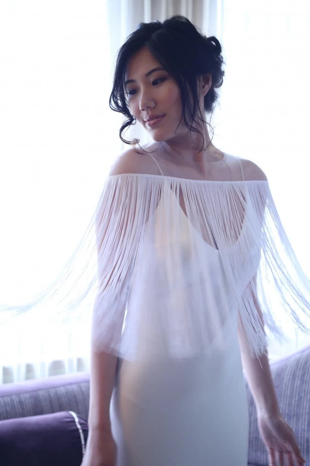 Bride in a fringed cape beside a bright window.