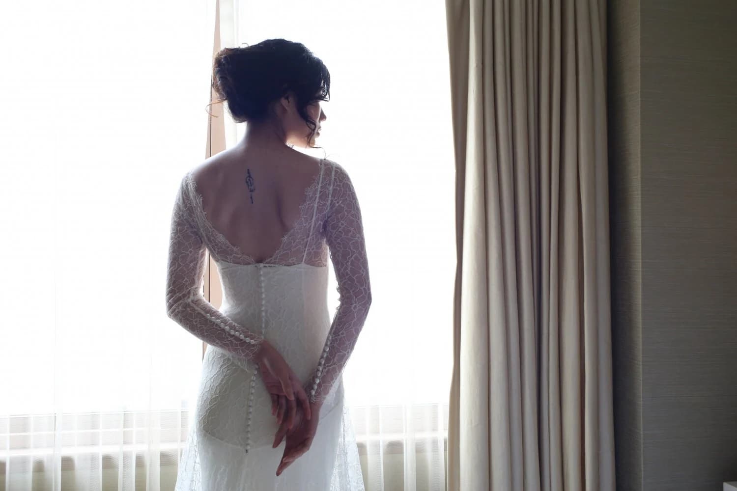 Bride viewed from behind in a lace gown by a window.