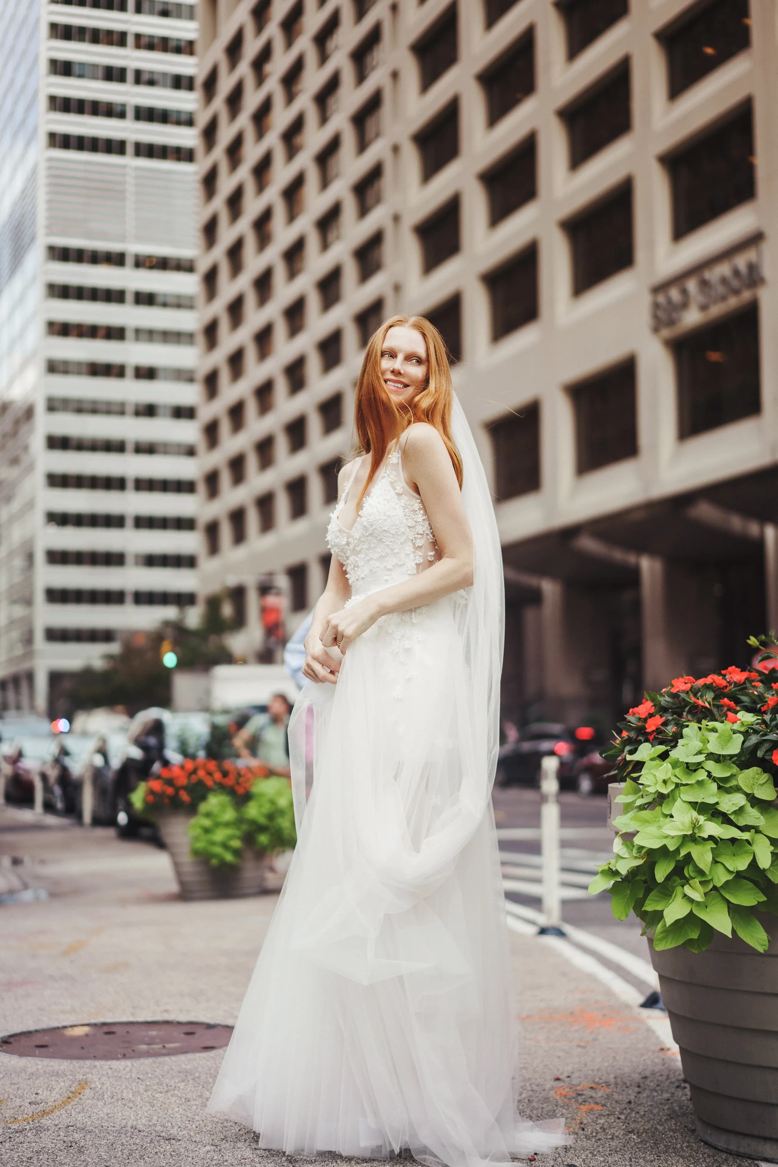 Bride in a veil and lace gown between tall city buildings.