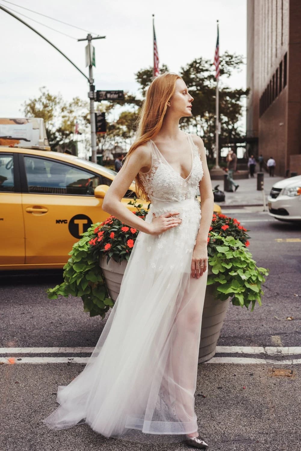 Bride in a white gown beside a yellow taxi and city planters.
