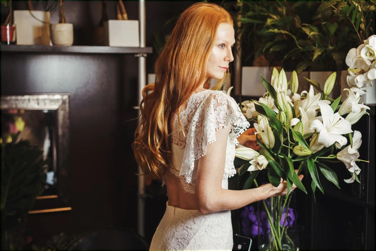Bride arranging white lilies while wearing a lace gown.