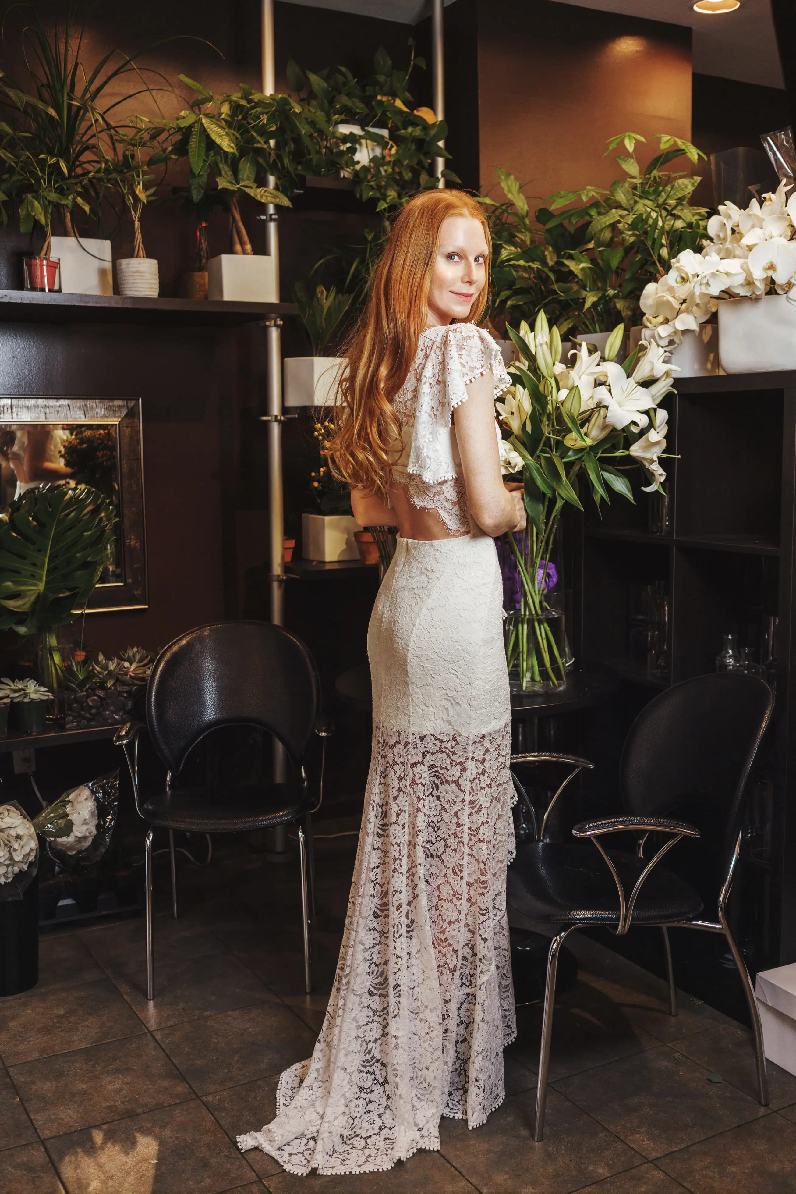Bride in a long lace gown beside lilies and indoor plants.