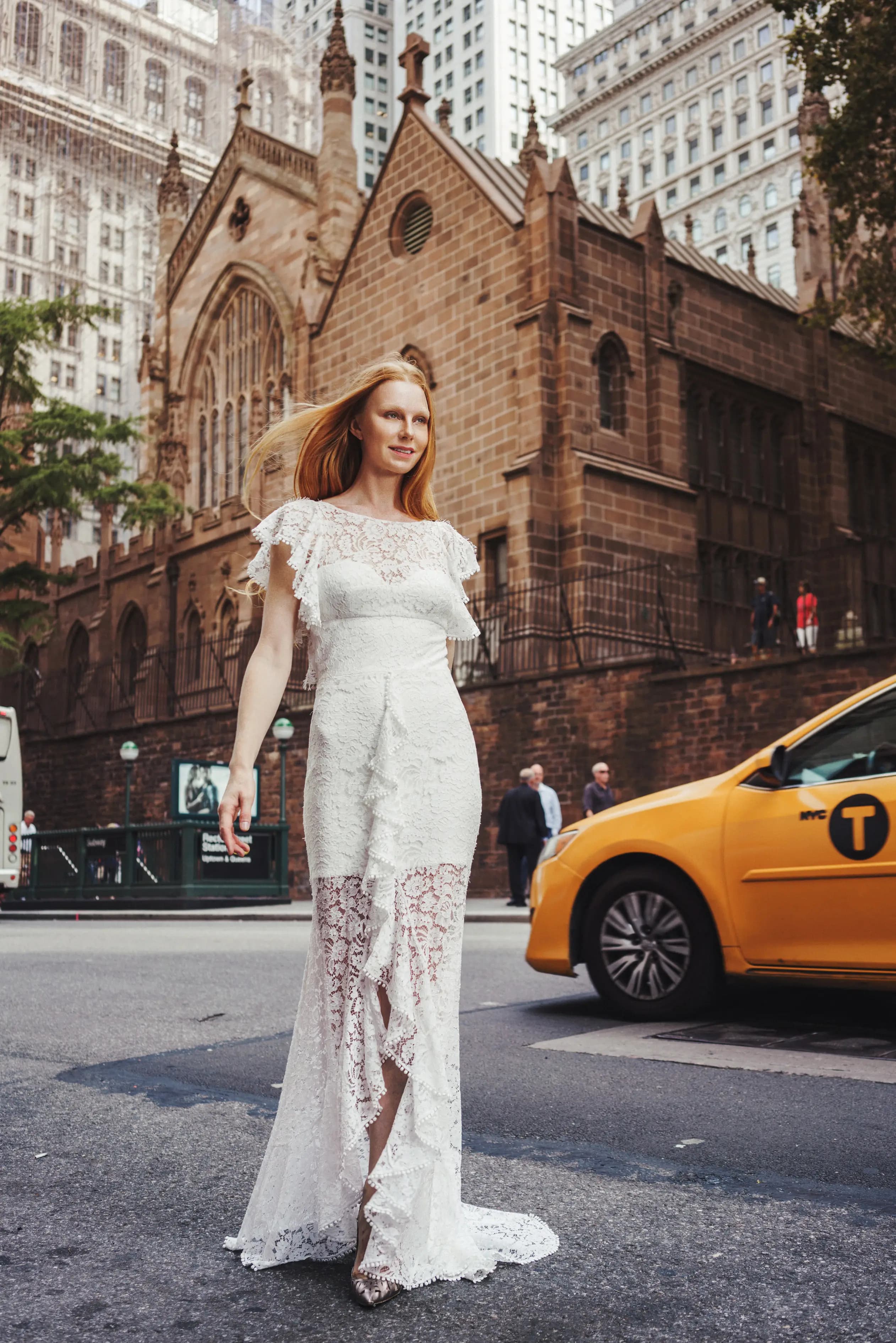 Bride in a floral lace gown beside a yellow taxi.