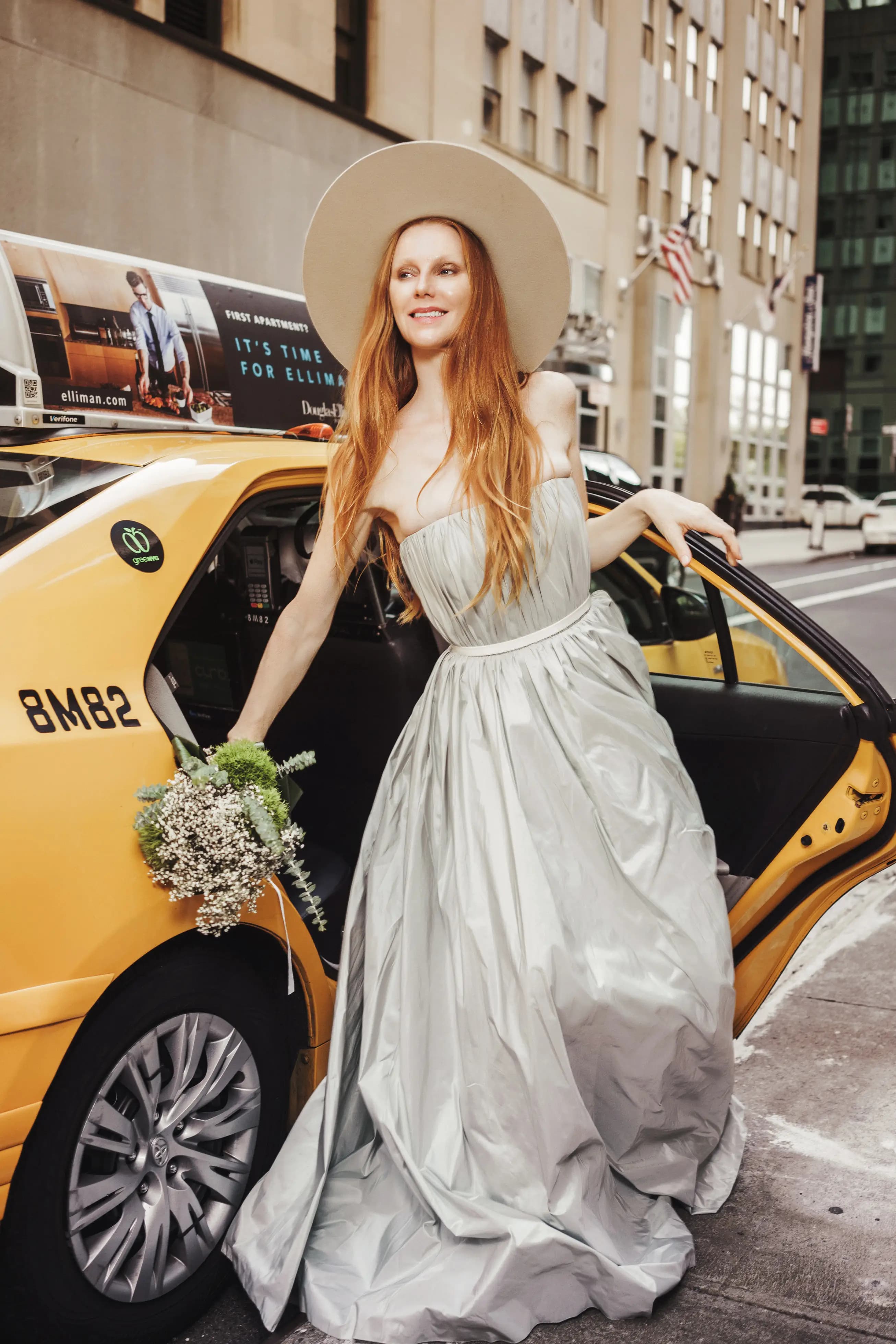 Bride in a wide-brim hat holding a bouquet beside a taxi.