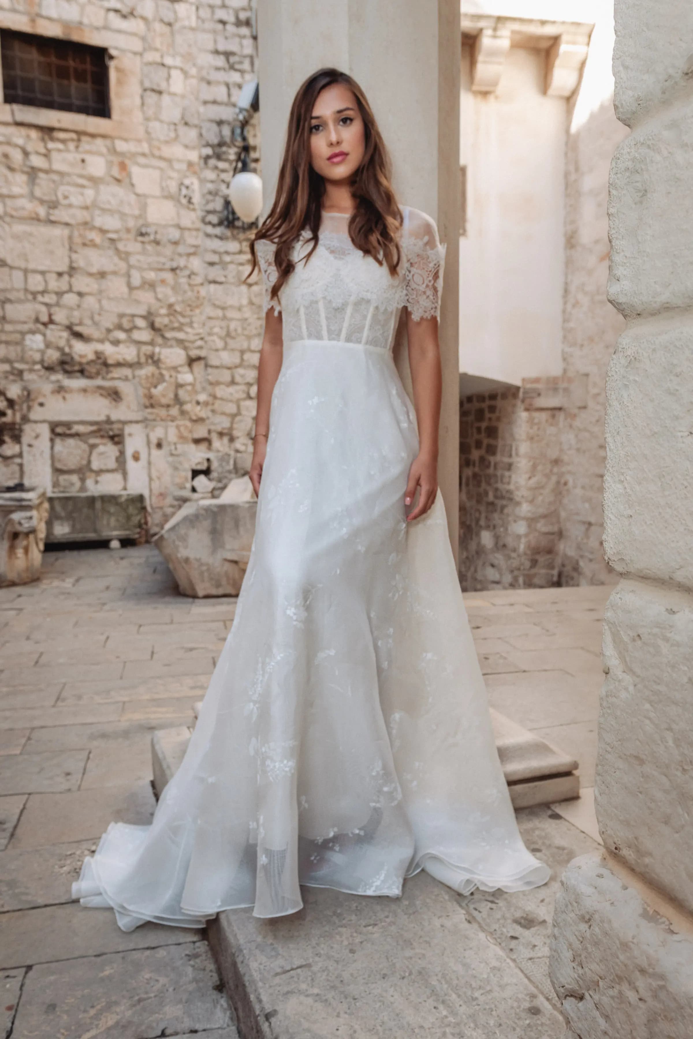 Bride in a lace gown between pale stone walls.
