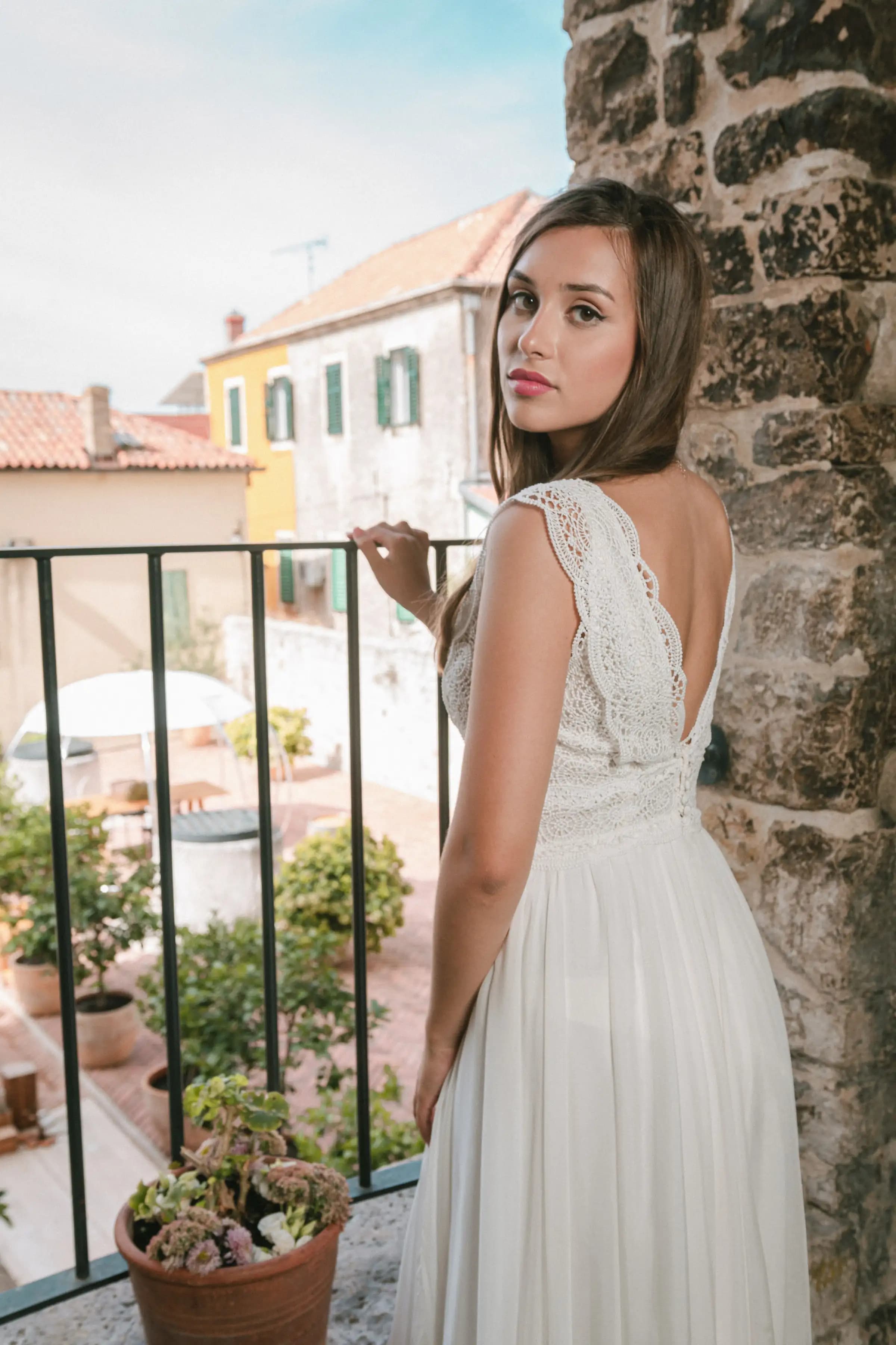 Bride showing the back of a lace gown beside a stone balcony.