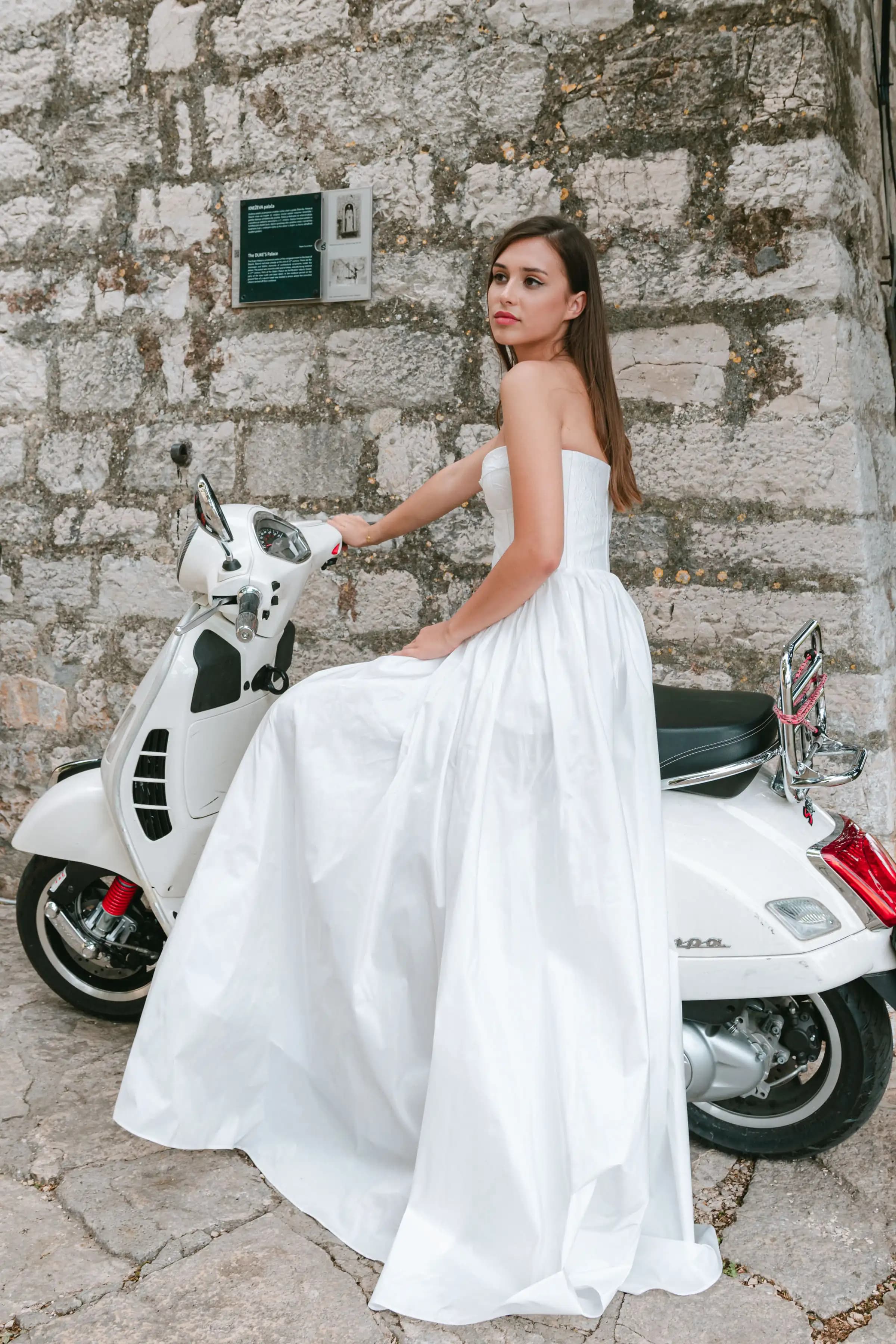 Bride seated on a white scooter beside a stone wall.